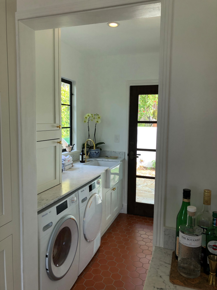 This is an example of a small mediterranean single-wall dedicated laundry room in Santa Barbara with a farmhouse sink, shaker cabinets, white cabinets, marble benchtops, white splashback, marble splashback, white walls, terra-cotta floors, a side-by-side washer and dryer, red floor and white benchtop.