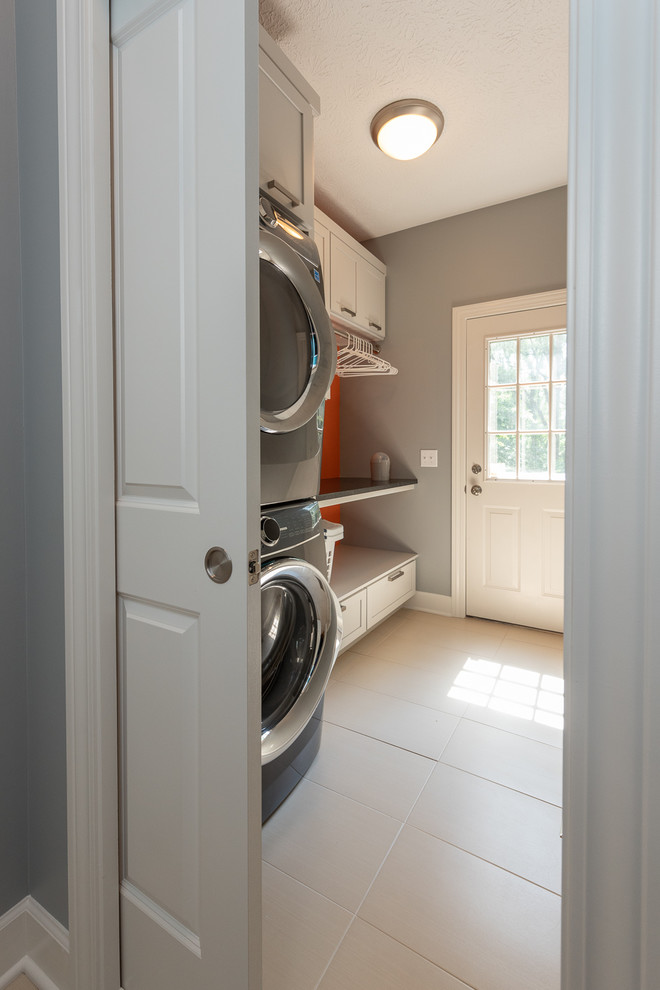 This is an example of a small traditional single-wall utility room in Indianapolis with recessed-panel cabinets, grey cabinets, granite benchtops, grey walls, ceramic floors, a stacked washer and dryer, white floor and black benchtop.