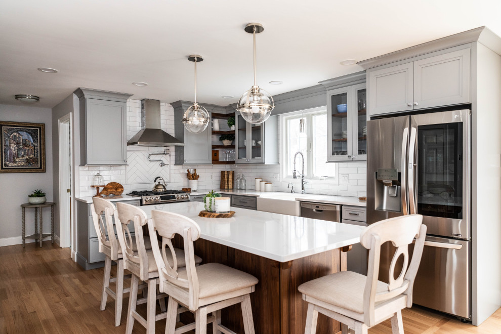 Inredning av ett klassiskt vit vitt l-kök, med en rustik diskho, luckor med infälld panel, grå skåp, vitt stänkskydd, rostfria vitvaror, mellanmörkt trägolv, en köksö och brunt golv