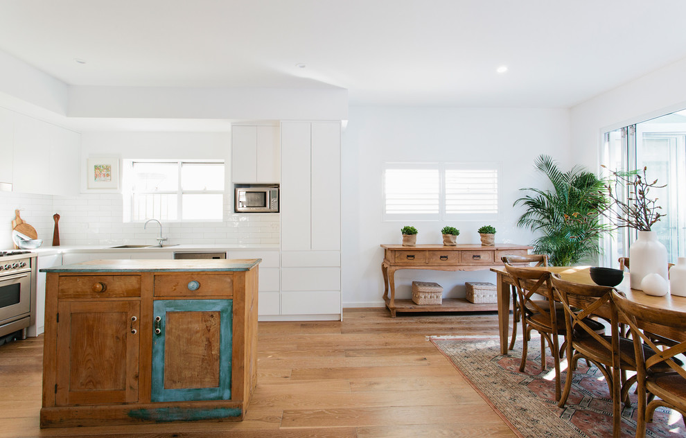 Inspiration for a large beach style open plan kitchen in Sydney with a double-bowl sink, flat-panel cabinets, white cabinets, white splashback, subway tile splashback, stainless steel appliances, light hardwood floors and with island.