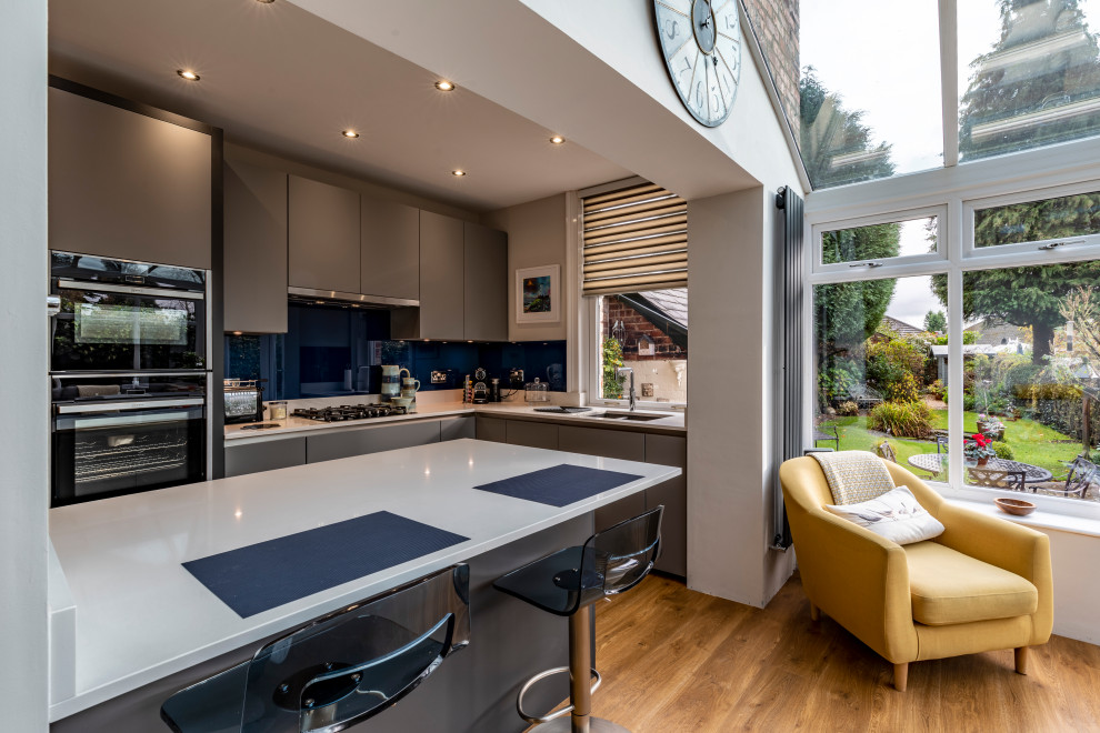 Design ideas for a small modern l-shaped open plan kitchen in Manchester with a double-bowl sink, flat-panel cabinets, grey cabinets, blue splashback, glass sheet splashback, black appliances, light hardwood floors, with island and grey benchtop.