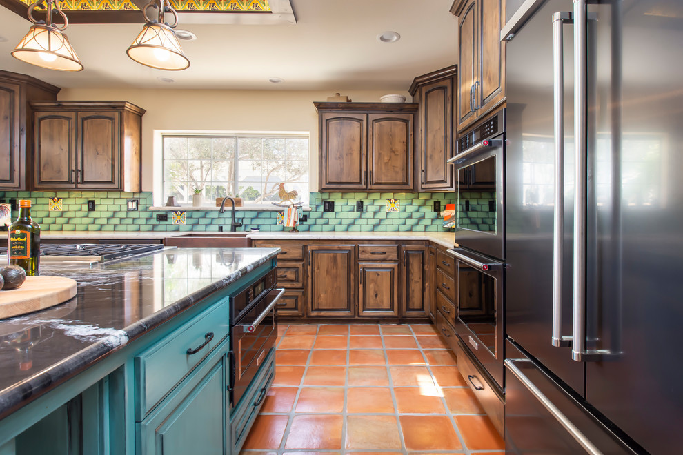 This is an example of a large u-shaped eat-in kitchen in Los Angeles with a farmhouse sink, raised-panel cabinets, medium wood cabinets, quartz benchtops, green splashback, ceramic splashback, stainless steel appliances, terra-cotta floors, with island, orange floor and black benchtop.