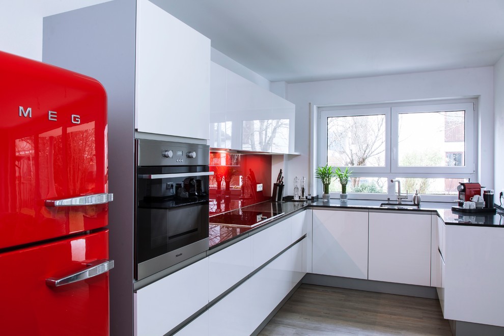 Mid-sized contemporary u-shaped kitchen in Munich with an integrated sink, flat-panel cabinets, white cabinets, red splashback, glass sheet splashback, coloured appliances, no island and light hardwood floors.