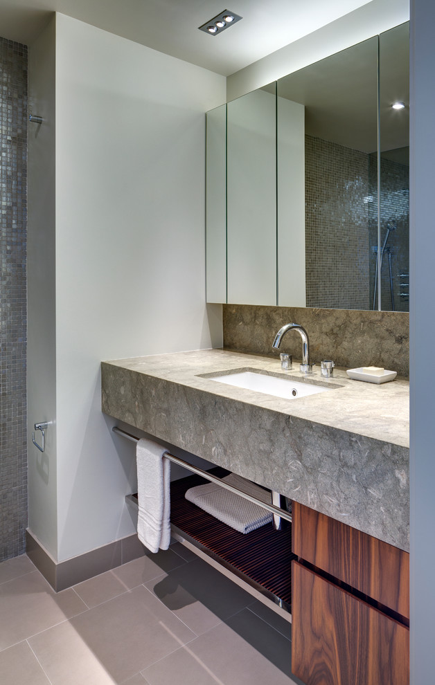 Photo of a small contemporary bathroom in New York with flat-panel cabinets, medium wood cabinets, gray tile, an undermount sink, limestone benchtops, an alcove shower, a one-piece toilet, porcelain floors and limestone.