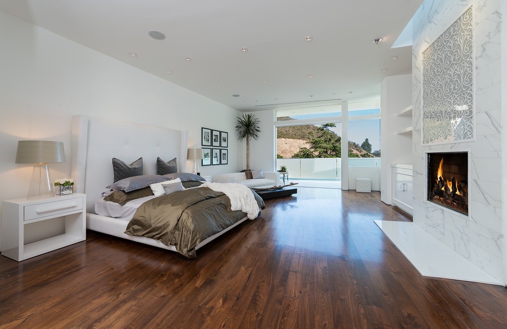 Photo of a contemporary master bedroom in Los Angeles with white walls, dark hardwood floors, a standard fireplace, a stone fireplace surround and brown floor.
