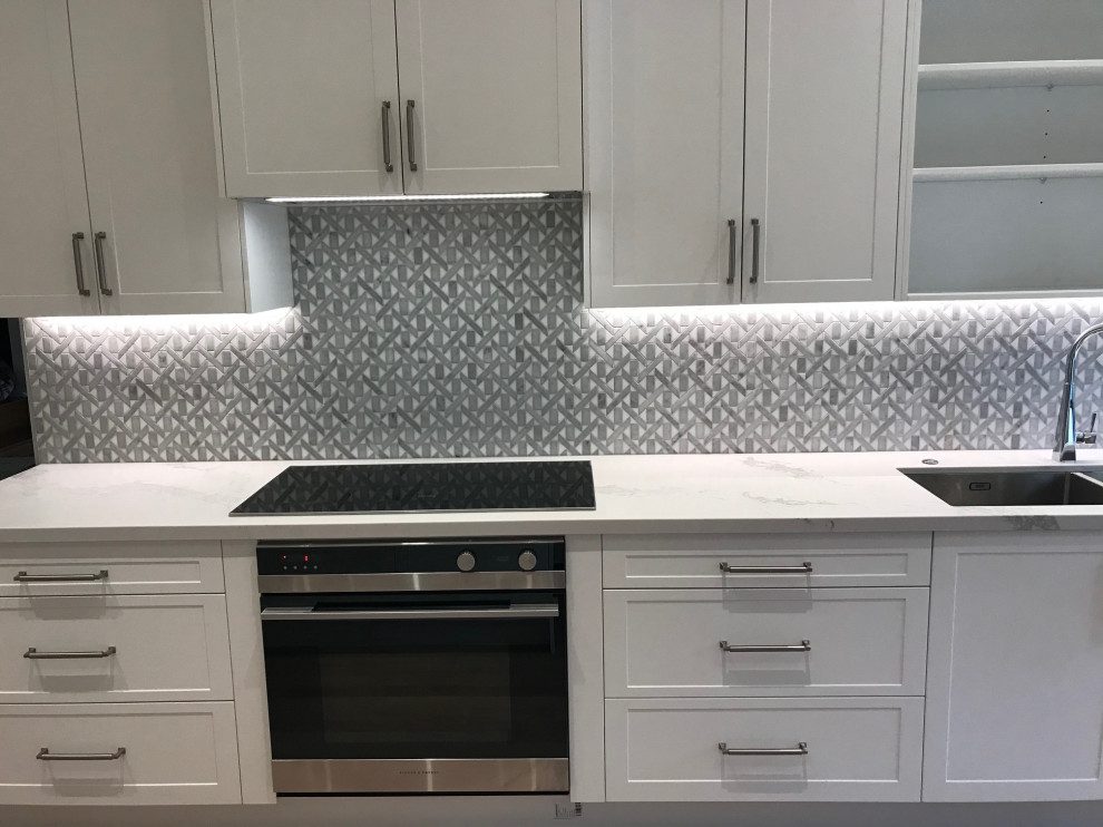 This is an example of a large modern l-shaped kitchen pantry in Sydney with an undermount sink, shaker cabinets, white cabinets, quartz benchtops, grey splashback, mosaic tile splashback, black appliances, medium hardwood floors, with island, brown floor and white benchtop.