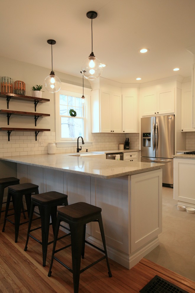 Example of a mid-sized transitional l-shaped porcelain tile and gray floor eat-in kitchen design in Cincinnati with a farmhouse sink, shaker cabinets, white cabinets, quartz countertops, white backsplash, subway tile backsplash, stainless steel appliances and an island