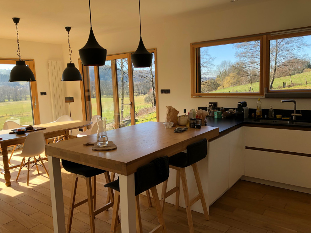 This is an example of a mid-sized modern u-shaped open plan kitchen in Nancy with an undermount sink, flat-panel cabinets, light wood cabinets, wood benchtops, black splashback, granite splashback, black appliances, light hardwood floors, with island, beige floor and beige benchtop.
