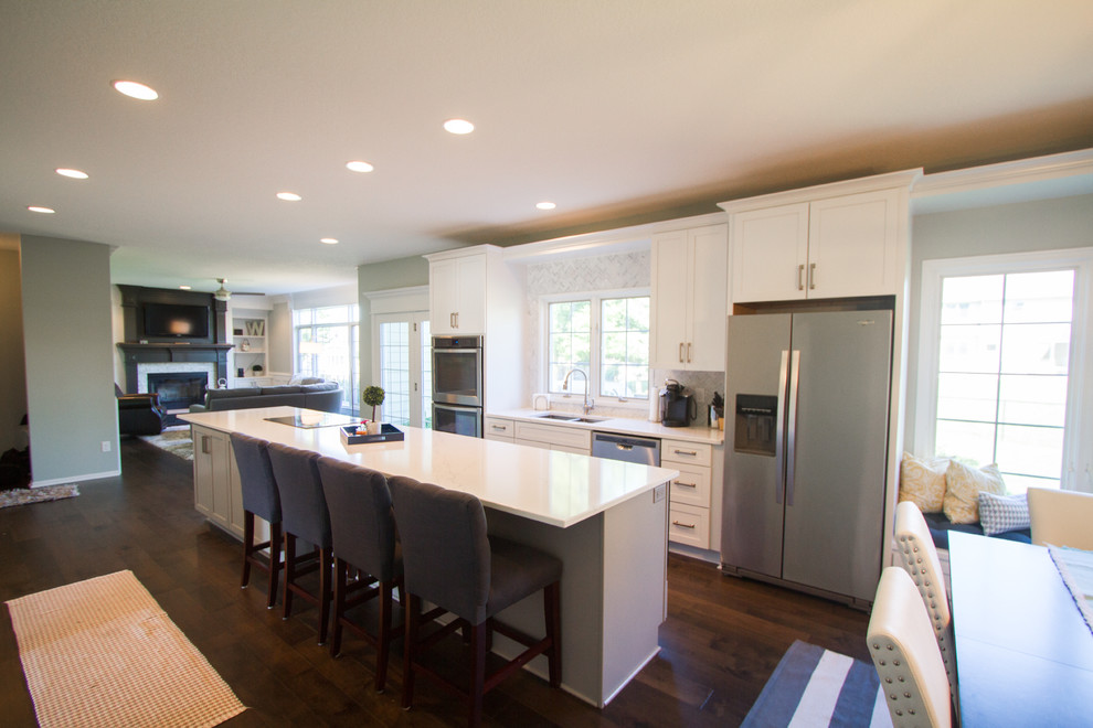 Example of a transitional single-wall dark wood floor and brown floor open concept kitchen design with an undermount sink, recessed-panel cabinets, white cabinets, quartzite countertops, gray backsplash, marble backsplash, stainless steel appliances and an island