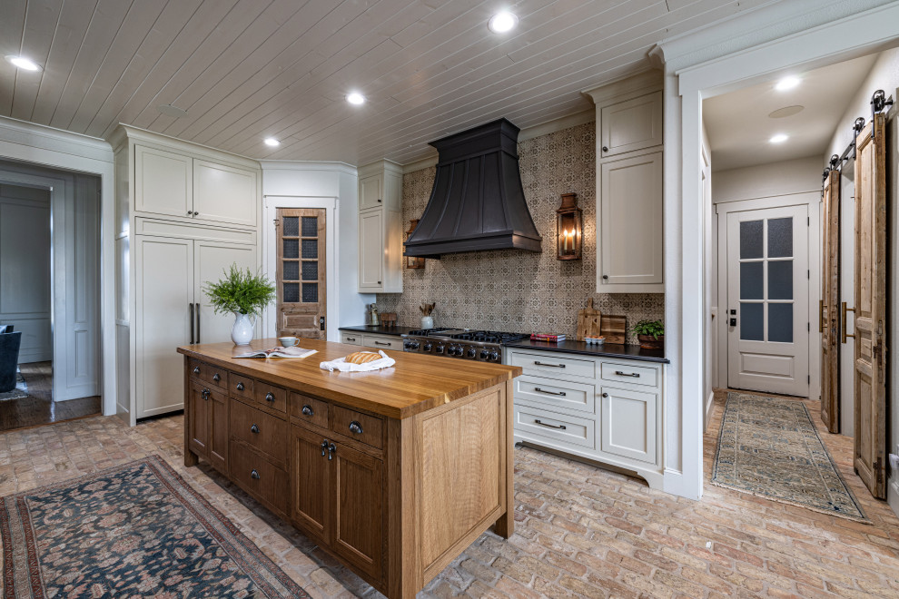 Photo of a large country u-shaped eat-in kitchen in Dallas with a farmhouse sink, beaded inset cabinets, beige cabinets, granite benchtops, beige splashback, terra-cotta splashback, panelled appliances, brick floors, brown floor, black benchtop, timber and multiple islands.