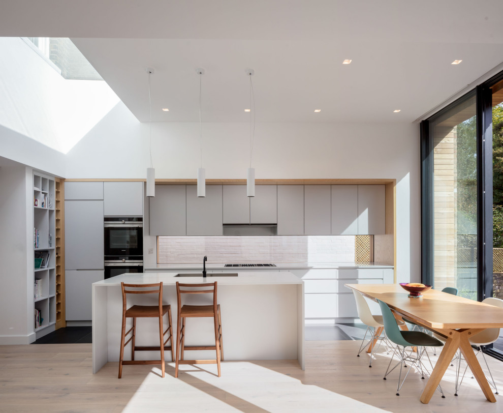 Large modern l-shaped open plan kitchen in London with an integrated sink, flat-panel cabinets, grey cabinets, solid surface benchtops, grey splashback, stainless steel appliances, light hardwood floors, with island, beige floor and white benchtop.