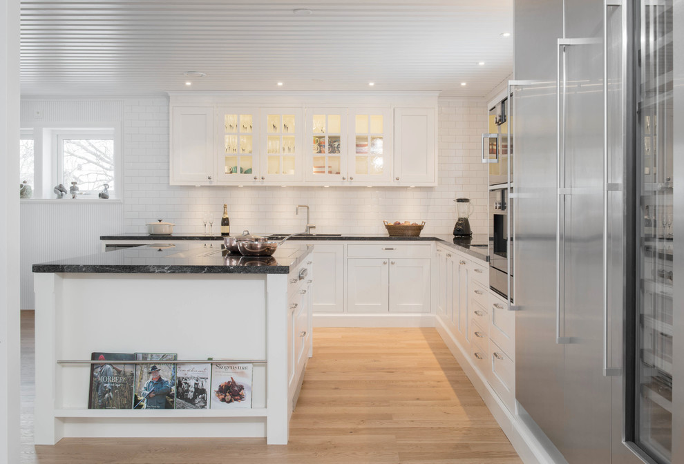 Design ideas for a large traditional l-shaped eat-in kitchen in Stockholm with an undermount sink, shaker cabinets, white cabinets, granite benchtops, white splashback, porcelain splashback, stainless steel appliances, light hardwood floors and with island.