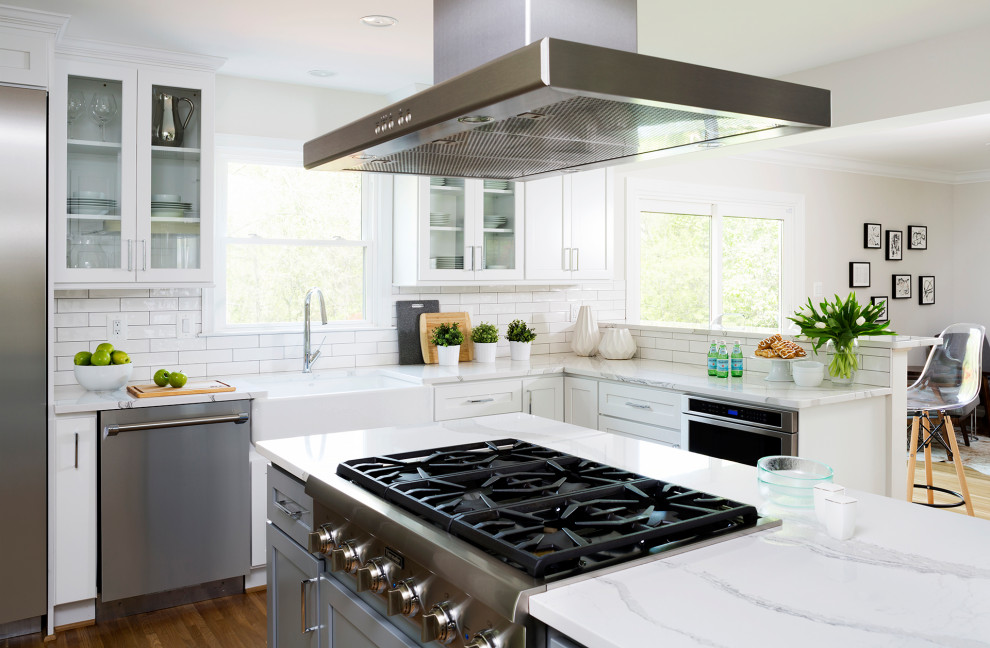 Transitional u-shaped medium tone wood floor and brown floor kitchen photo in DC Metro with a farmhouse sink, shaker cabinets, white cabinets, white backsplash, stainless steel appliances, an island and white countertops