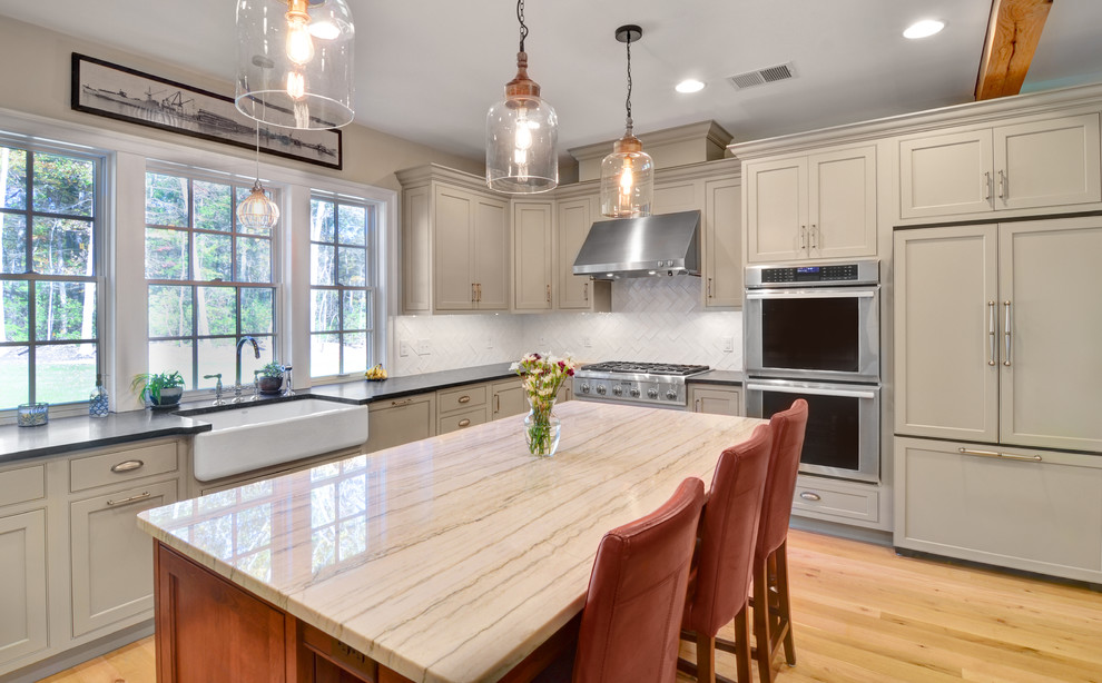 Photo of a large country u-shaped kitchen in Baltimore with a farmhouse sink, shaker cabinets, beige cabinets, soapstone benchtops, white splashback, glass tile splashback, panelled appliances, light hardwood floors, with island and brown floor.