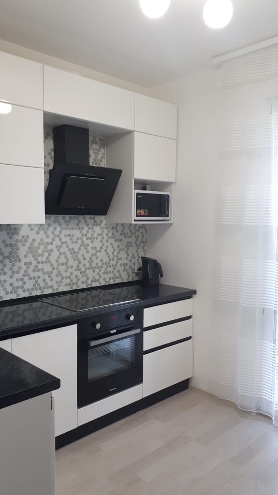 This is an example of a mid-sized contemporary l-shaped separate kitchen in Saint Petersburg with a drop-in sink, flat-panel cabinets, white cabinets, wood benchtops, grey splashback, mosaic tile splashback, black appliances, laminate floors, no island, beige floor and black benchtop.