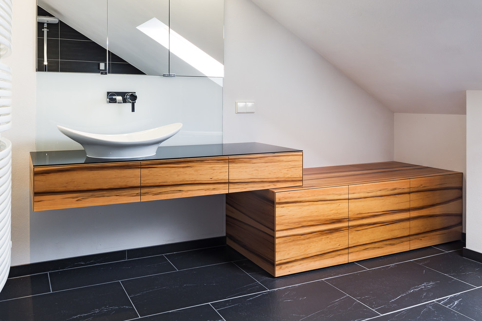 Photo of a small contemporary master bathroom in Other with flat-panel cabinets, medium wood cabinets, a freestanding tub, a curbless shower, a wall-mount toilet, white tile, glass sheet wall, white walls, slate floors, a vessel sink, glass benchtops, black floor, an open shower and black benchtops.