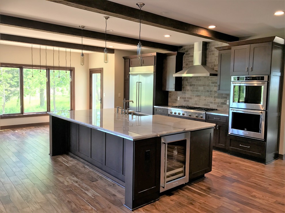 This is an example of a large transitional single-wall open plan kitchen in Milwaukee with a farmhouse sink, shaker cabinets, dark wood cabinets, grey splashback, stainless steel appliances, dark hardwood floors, with island, brown floor, onyx benchtops and brick splashback.
