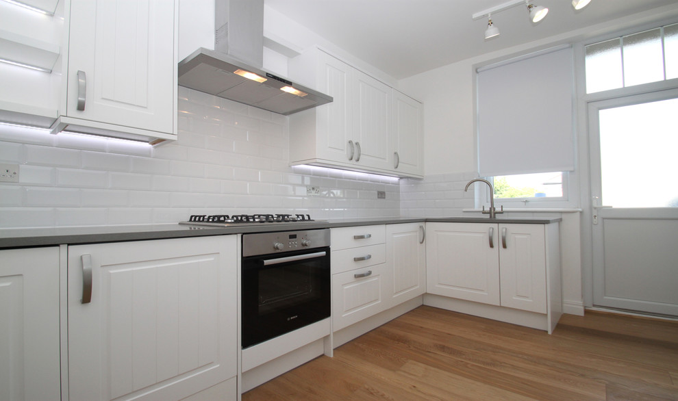 This is an example of a small traditional l-shaped separate kitchen in London with a drop-in sink, raised-panel cabinets, grey cabinets, laminate benchtops, white splashback, ceramic splashback, stainless steel appliances, medium hardwood floors, a peninsula and brown floor.