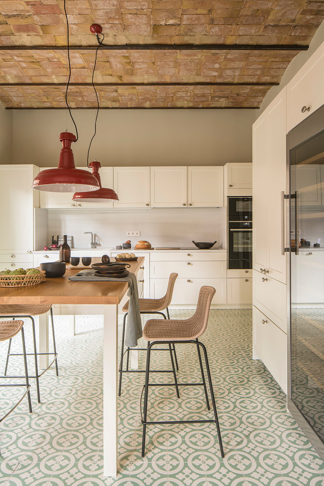 This is an example of a mid-sized mediterranean single-wall eat-in kitchen in Barcelona with an integrated sink, white cabinets, wood benchtops, white splashback, limestone splashback, with island, brown benchtop, shaker cabinets, black appliances and green floor.