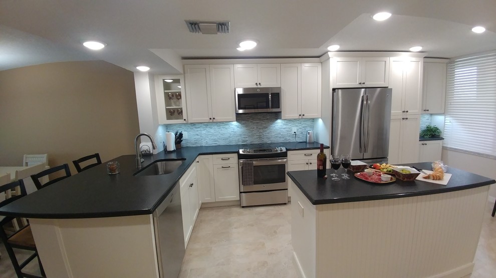 Photo of a mid-sized tropical l-shaped open plan kitchen in Tampa with a single-bowl sink, shaker cabinets, white cabinets, granite benchtops, multi-coloured splashback, mosaic tile splashback, stainless steel appliances, porcelain floors, with island and beige floor.