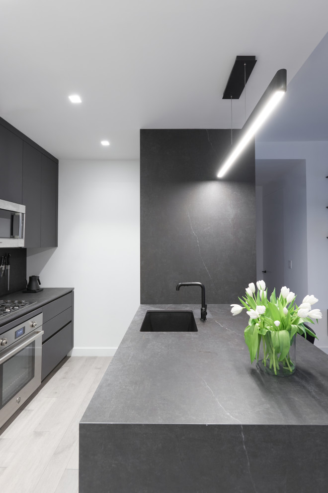 Photo of a small contemporary galley kitchen in New York with flat-panel cabinets, black cabinets, solid surface benchtops, black splashback, stainless steel appliances, medium hardwood floors, with island and grey benchtop.