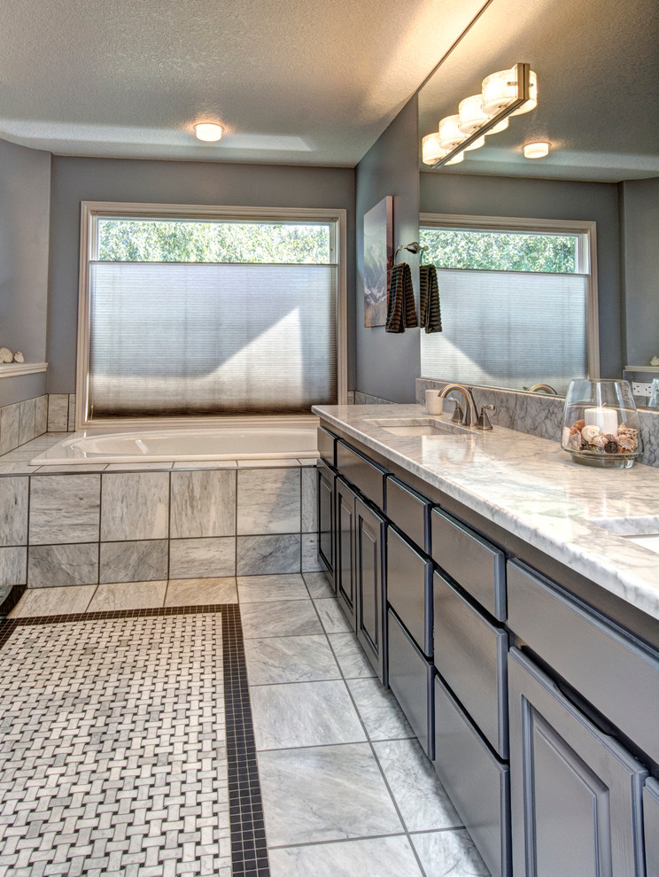 This is an example of a large transitional master bathroom in Portland with an undermount sink, grey cabinets, marble benchtops, a drop-in tub, an alcove shower, a one-piece toilet, stone tile, marble floors, raised-panel cabinets, gray tile, grey walls, grey floor and a hinged shower door.