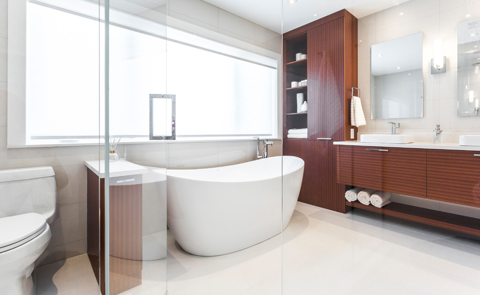 This is an example of a mid-sized modern master bathroom in Montreal with flat-panel cabinets, medium wood cabinets, a freestanding tub, a double shower, a one-piece toilet, white tile, ceramic tile, white walls, porcelain floors, a vessel sink, quartzite benchtops, beige floor, a sliding shower screen and white benchtops.