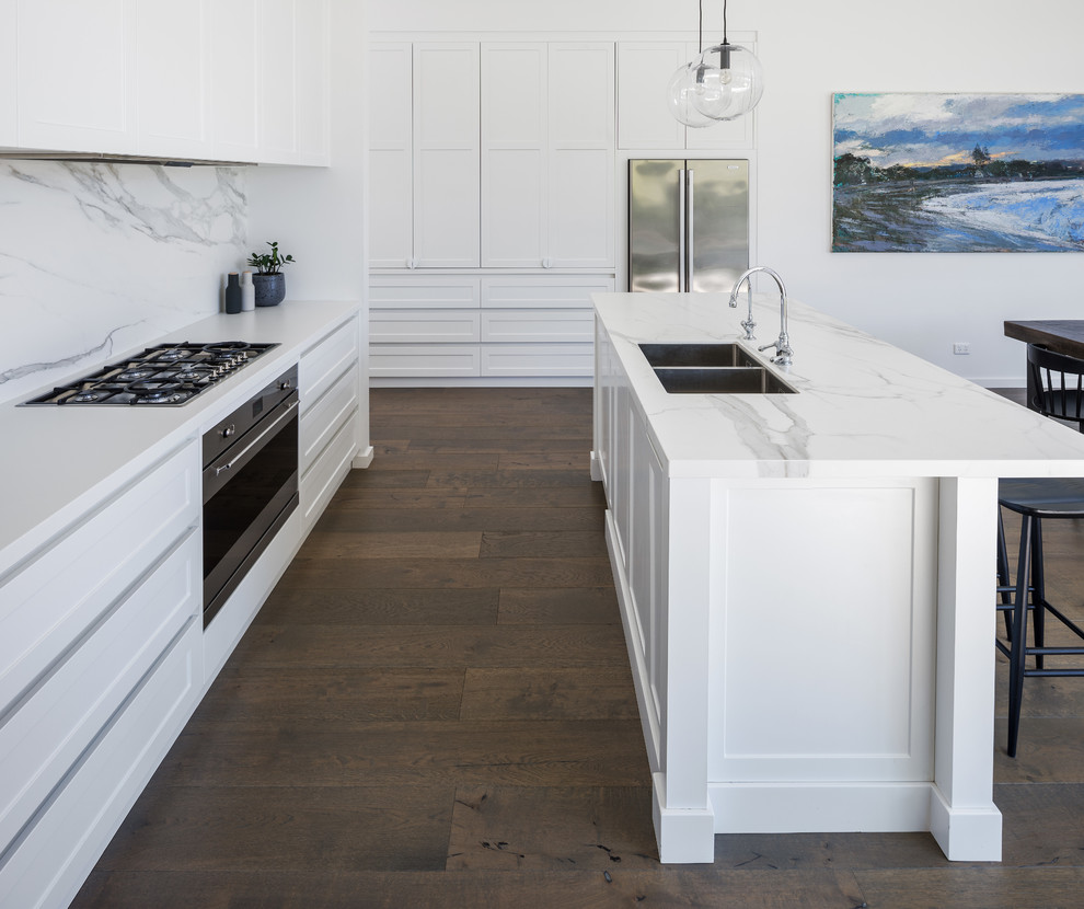 This is an example of a contemporary galley open plan kitchen in Perth with an undermount sink, shaker cabinets, white cabinets, quartz benchtops, white splashback, marble splashback, stainless steel appliances, dark hardwood floors, with island and white benchtop.