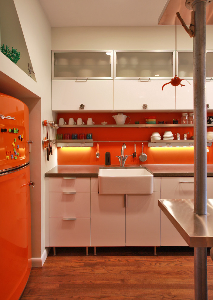 Photo of a small eclectic galley separate kitchen in DC Metro with a farmhouse sink, flat-panel cabinets, white cabinets, concrete benchtops, orange splashback, stainless steel appliances, medium hardwood floors and brown floor.