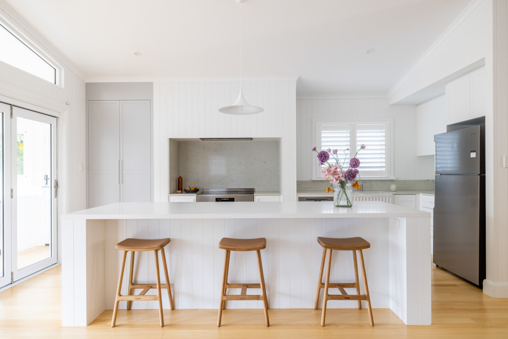 Mid-sized beach style l-shaped eat-in kitchen in Townsville with shaker cabinets, white cabinets, quartz benchtops, light hardwood floors, with island and white benchtop.