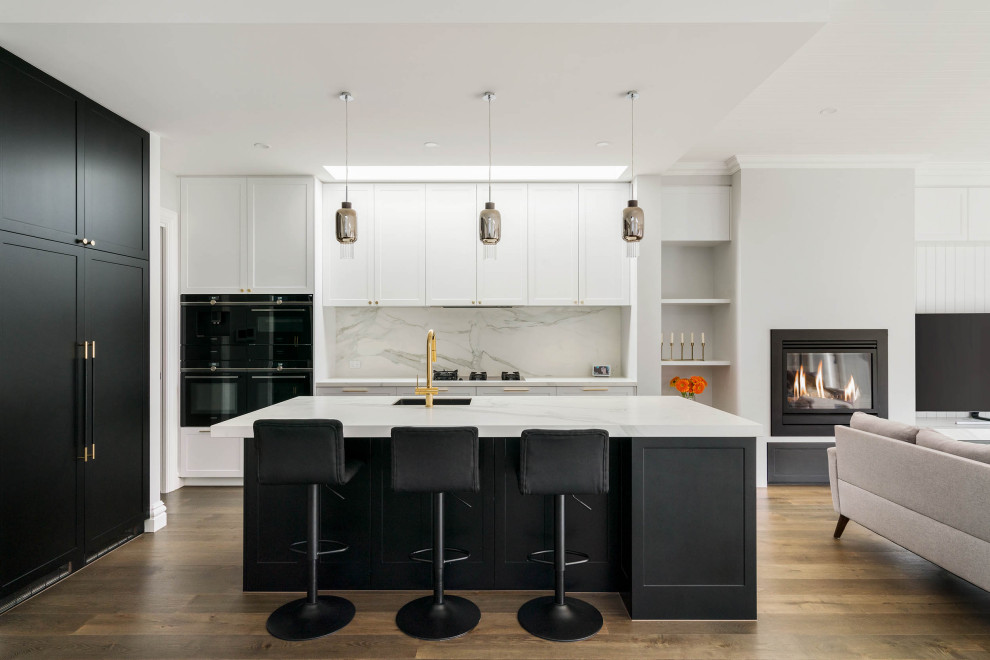 Inspiration for a transitional l-shaped open plan kitchen in Melbourne with an undermount sink, recessed-panel cabinets, white cabinets, white splashback, stone slab splashback, black appliances, medium hardwood floors, with island, brown floor and white benchtop.