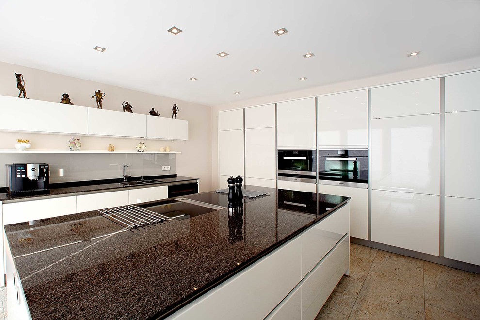 Inspiration for a large contemporary galley open plan kitchen in Frankfurt with a double-bowl sink, flat-panel cabinets, white cabinets, grey splashback, glass sheet splashback, with island, marble benchtops, black appliances and travertine floors.
