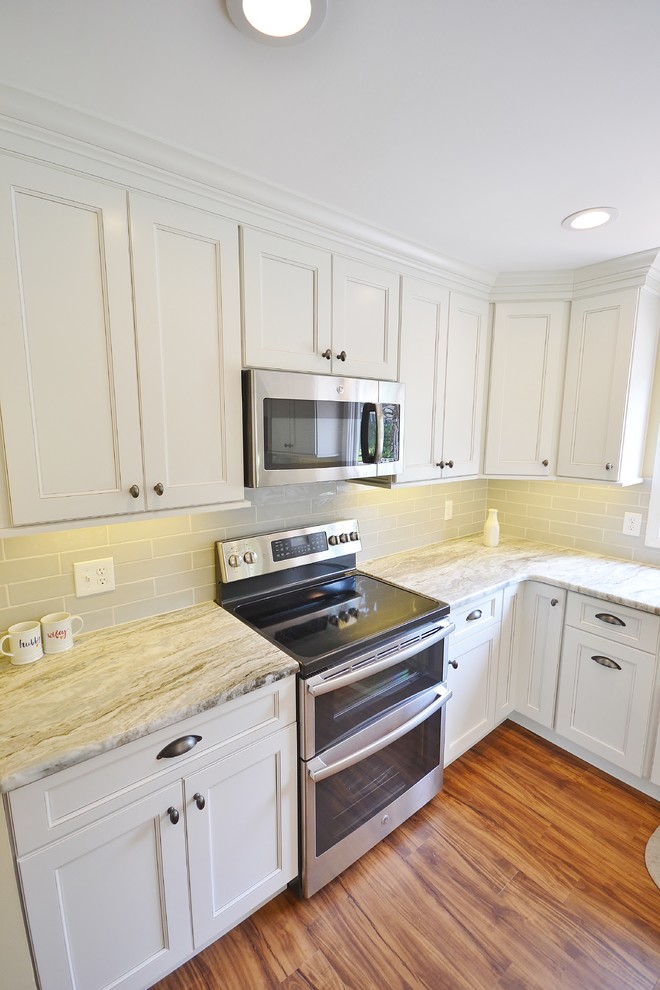 This is an example of a mid-sized transitional l-shaped eat-in kitchen in Philadelphia with a farmhouse sink, raised-panel cabinets, white cabinets, quartzite benchtops, beige splashback, subway tile splashback, stainless steel appliances, dark hardwood floors and with island.