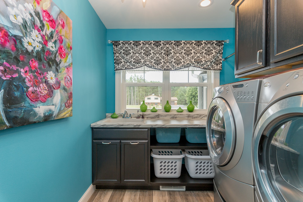 Transitional l-shaped dedicated laundry room in Other with a drop-in sink, recessed-panel cabinets, black cabinets, blue walls, light hardwood floors, a side-by-side washer and dryer, beige floor and grey benchtop.