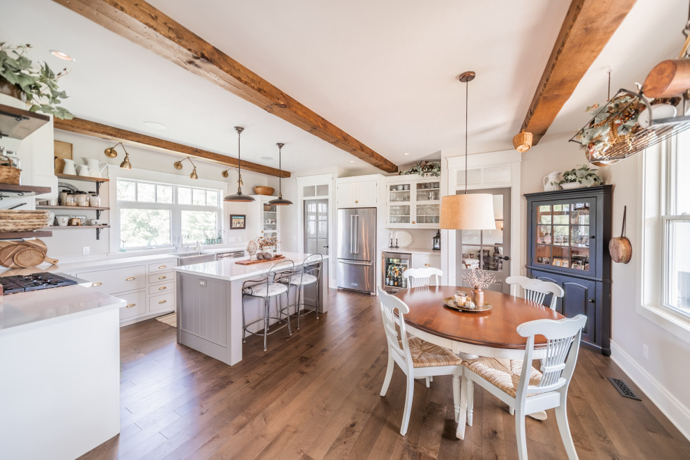 Large traditional l-shaped eat-in kitchen with a farmhouse sink, flat-panel cabinets, white cabinets, quartz benchtops, beige splashback, stainless steel appliances, medium hardwood floors, with island, brown floor and white benchtop.