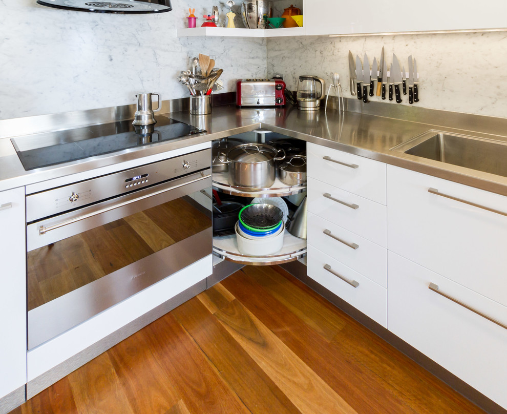 Photo of a small contemporary l-shaped kitchen pantry in Melbourne with an integrated sink, flat-panel cabinets, white cabinets, stainless steel benchtops, white splashback, marble splashback, stainless steel appliances, medium hardwood floors and brown floor.