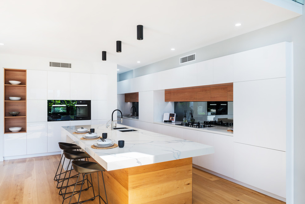 This is an example of a contemporary l-shaped kitchen in Brisbane with an undermount sink, flat-panel cabinets, white cabinets, black appliances, medium hardwood floors, with island, brown floor, white benchtop and marble benchtops.