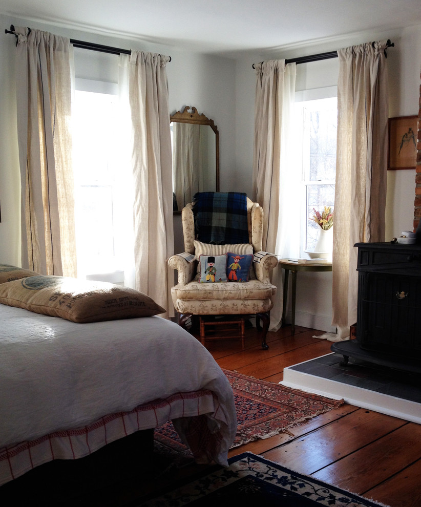 This is an example of a mid-sized country guest bedroom in New York with white walls, medium hardwood floors, a wood stove and a brick fireplace surround.