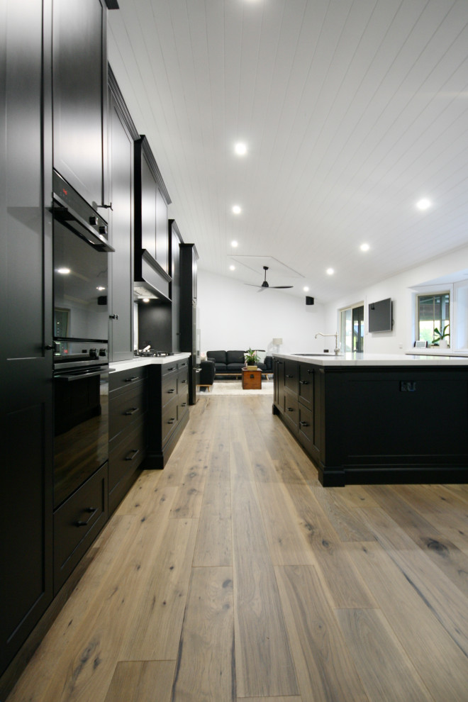 Design ideas for an expansive classic galley open plan kitchen in Sydney with a submerged sink, shaker cabinets, black cabinets, engineered stone countertops, white splashback, engineered quartz splashback, stainless steel appliances, medium hardwood flooring, an island, brown floors and white worktops.