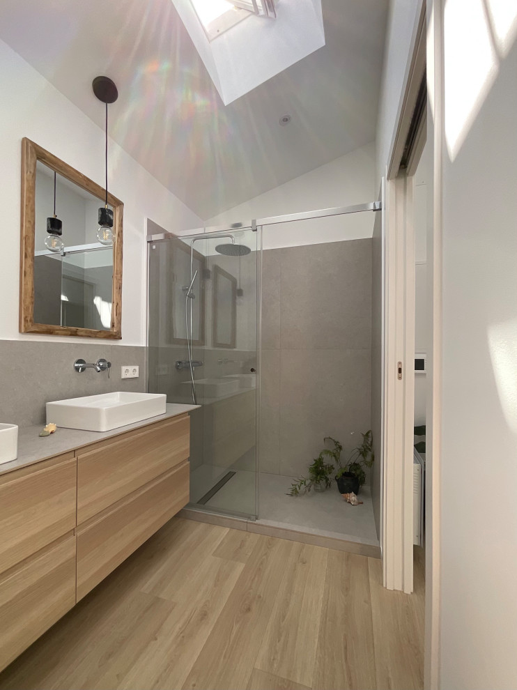 This is an example of a mid-sized modern master bathroom in Barcelona with white cabinets, a curbless shower, a wall-mount toilet, gray tile, white walls, laminate floors, a vessel sink, tile benchtops, brown floor, a sliding shower screen, grey benchtops, a double vanity and a floating vanity.