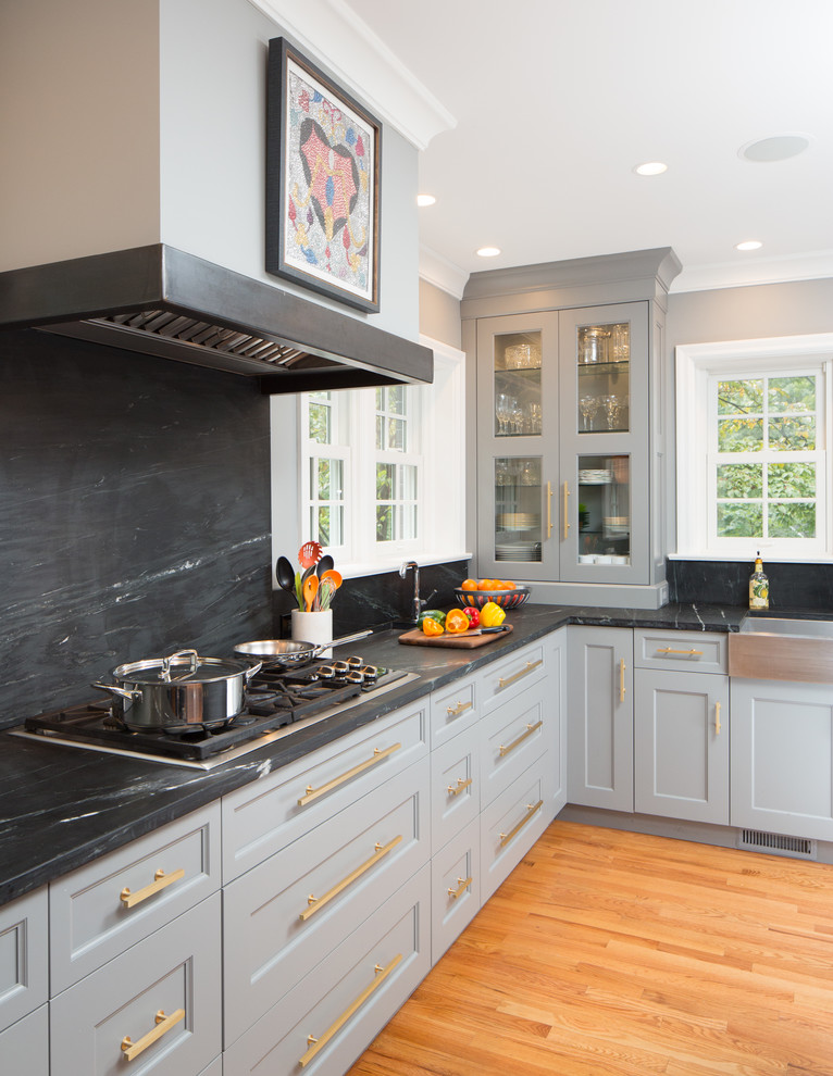Inspiration for a mid-sized transitional l-shaped open plan kitchen in St Louis with a farmhouse sink, recessed-panel cabinets, grey cabinets, soapstone benchtops, stone slab splashback, panelled appliances, medium hardwood floors and with island.