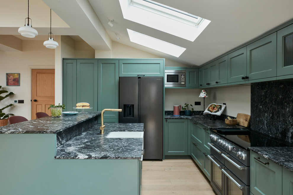 Example of a mid-sized classic l-shaped light wood floor and vaulted ceiling open concept kitchen design in London with a farmhouse sink, recessed-panel cabinets, green cabinets, granite countertops, black backsplash, granite backsplash, an island and black countertops
