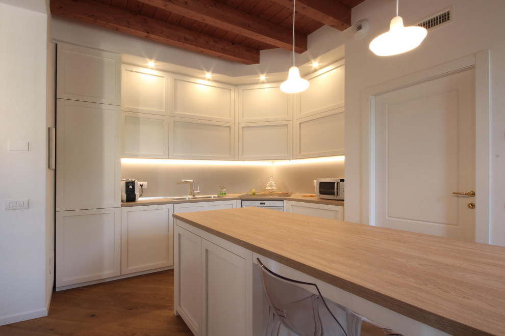 This is an example of a large country l-shaped open plan kitchen in Milan with a drop-in sink, recessed-panel cabinets, white cabinets, wood benchtops, grey splashback, slate splashback, white appliances, medium hardwood floors, with island, brown floor, brown benchtop and exposed beam.