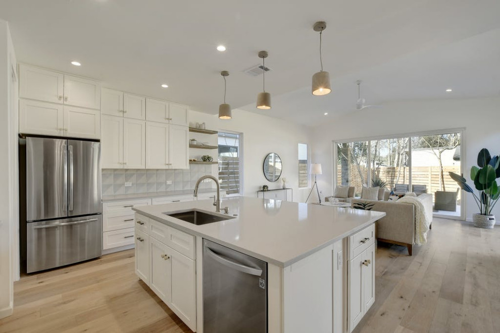 This is an example of a retro l-shaped open plan kitchen in Austin with a submerged sink, shaker cabinets, white cabinets, grey splashback, stainless steel appliances, medium hardwood flooring and an island.