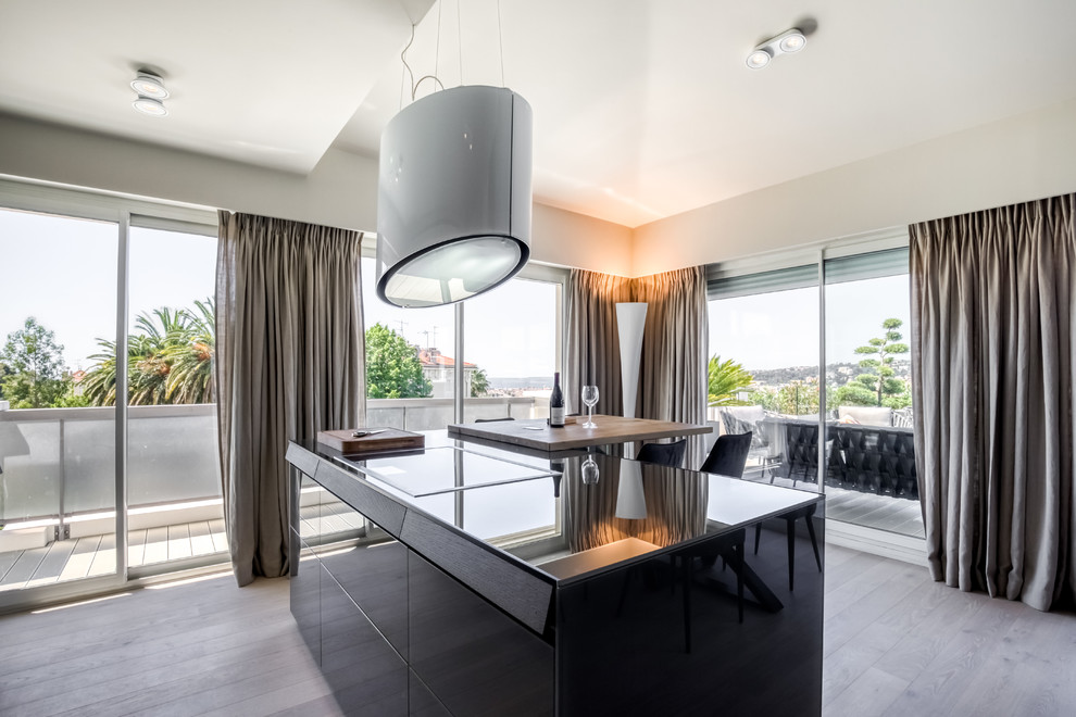 Photo of a large contemporary galley eat-in kitchen in Nice with an undermount sink, glass benchtops, white splashback, black appliances, light hardwood floors, with island and grey benchtop.