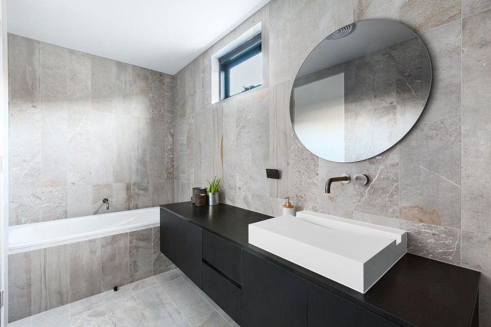 This is an example of a large contemporary kids bathroom in Melbourne with flat-panel cabinets, black cabinets, a drop-in tub, an alcove shower, a one-piece toilet, gray tile, marble, grey walls, marble floors, a console sink, marble benchtops, grey floor, an open shower and black benchtops.