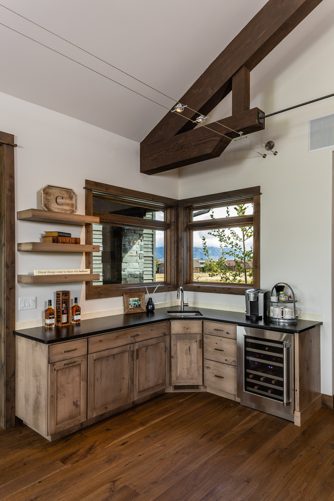 Photo of a country l-shaped wet bar in Other with an undermount sink, brown floor, black benchtop, light wood cabinets and medium hardwood floors.
