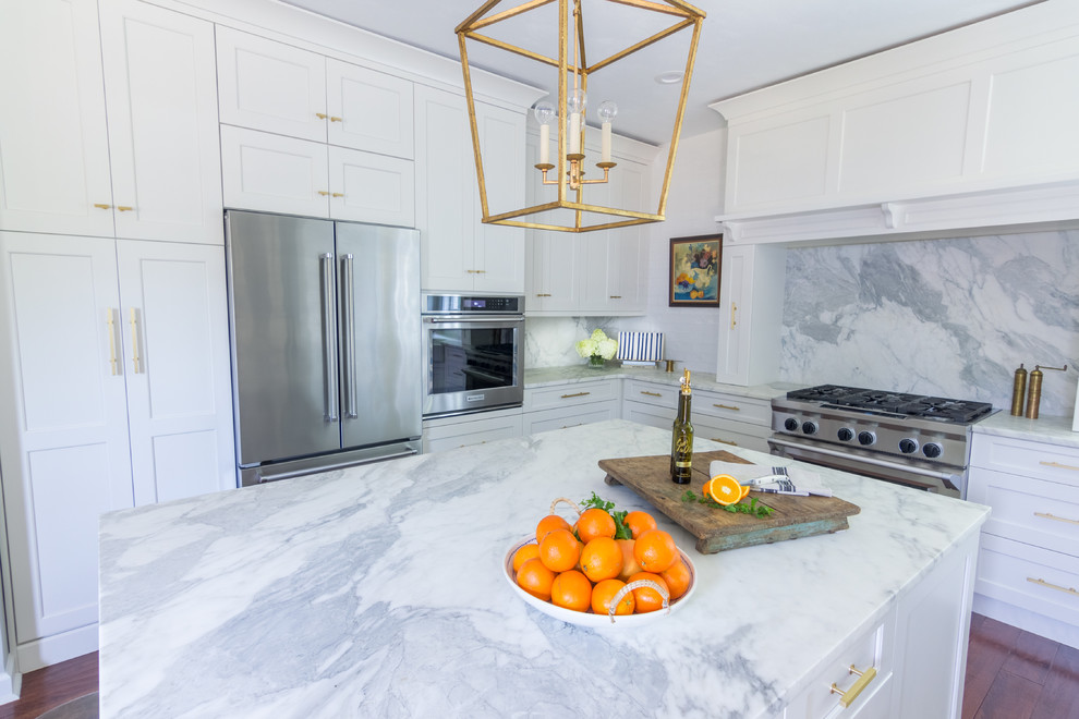 Example of a transitional u-shaped dark wood floor and brown floor kitchen design with a farmhouse sink, shaker cabinets, white cabinets, marble countertops, marble backsplash, stainless steel appliances and an island