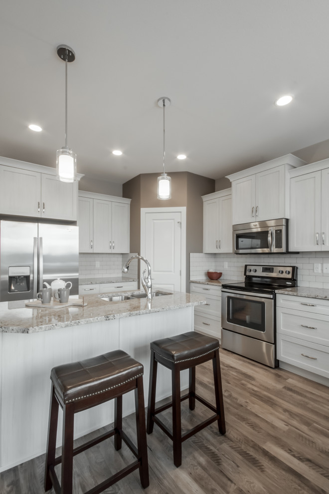 Mid-sized transitional l-shaped eat-in kitchen in Calgary with an undermount sink, shaker cabinets, white cabinets, granite benchtops, white splashback, subway tile splashback, stainless steel appliances, laminate floors and with island.