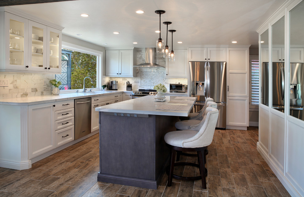 This is an example of a mid-sized transitional l-shaped open plan kitchen in Orange County with an undermount sink, recessed-panel cabinets, white cabinets, quartz benchtops, grey splashback, marble splashback, stainless steel appliances, porcelain floors, with island, brown floor and white benchtop.